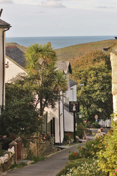 A narrow boscastle street with white houses, trees, and hills, leading to the distant sea.