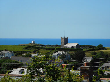 Boscastle coastal village with houses, a church, and green fields overlooking the blue sea under a clear sky.