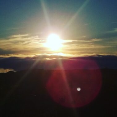 Bright sun setting over dark hills with clouds and vibrant lens flare in the evening sky.