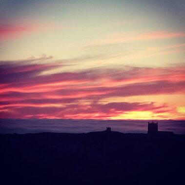 Silhouetted buildings beneath a vibrant sunset sky with streaks of pink, orange, and purple clouds.