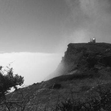 A small building stands on a cliff edge above clouds and grassy slopes in black and white.