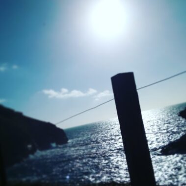 Blurred coastal scene with bright sun, ocean waves, and a silhouetted post in the foreground.