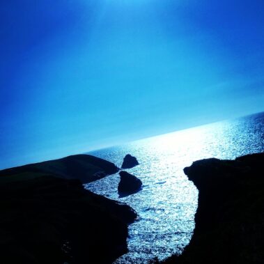 Rocky coastline and sea under a bright blue sky, with sunlight reflecting on the water.