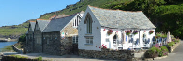 A charming white cottage café with outdoor seating by a river and hills in the background.